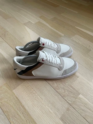 Vita Burberry sneakers med beige detaljer - Snygga vita sneakers från Burberry med beige mockadetaljer och klassiskt rutmönster på sidan. Skorna har vita snören, diskreta perforeringar och en låg silhuett. Perfekta för dig som gillar stilrena och trendiga skor med exklusiv känsla.