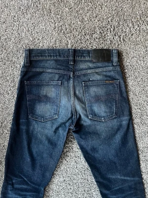 Mörkblå slim fit jeans från Nudie - Säljer ett par mörkblå slim fit jeans från Nudie med klassiska femficksdetaljer och läderpatch bak i midjan. Jeansen har en snygg tvätt med lätt slitning och raka ben. Perfekt för dig som gillar en stilren och modern look.