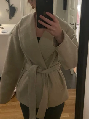 Beige kappa med knytskärp -  beige kappa med bred sjalkrage och knytskärp i midjan. En liten fläck på aemen