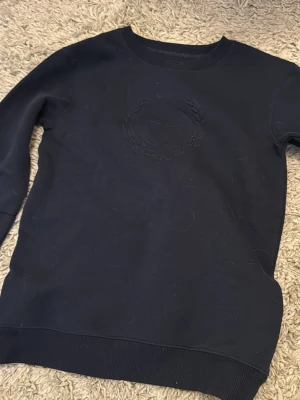 Svart stickad tröja med broderad logga - En svart stickad tröja med rund hals och ribbade muddar. Tröjan har en diskret broderad logga på bröstet i samma färg som tyget. Perfekt för dig som gillar en clean och stilren look.