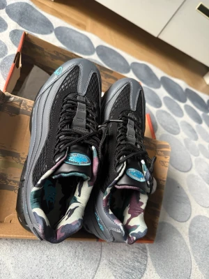 Nike Air Max 95 Corteiz grå/svart - Nike Air Max 95 Corteiz sneakers i grått och svart med blå detaljer. Ovandel i mesh och syntet, ikonisk Air Max-sula med blå luftbubblor. Fodret har camomönster i blått, lila och vitt. Snygg streetstil med broderad Corteiz-logga på sidan.