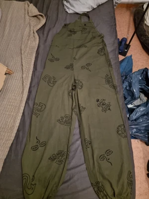 Grön jumpsuit med svarta mönster - Säljer en grön jumpsuit med svarta grafiska tryck och justerbara axelband. Jumpsuiten har avslappnad passform och resår vid bensluten. Perfekt för dig som gillar unika plagg med coola detaljer.