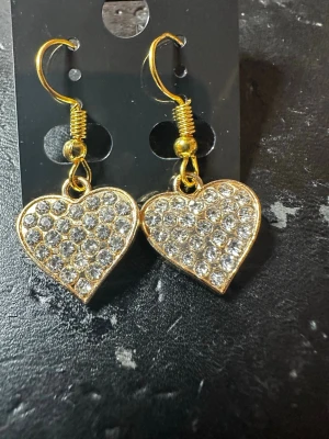 Guldiga hjärtörhängen med strass - Snygga örhängen i form av hjärtan med gnistrande klara strasstenar. Guldtonad metall och krokfäste. Perfekta för att ge din look lite extra bling och romantisk vibe.