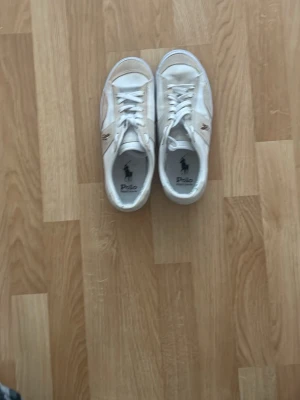 Vita sneakers från Polo Ralph Lauren - Säljer ett par klassiska vita sneakers från Polo Ralph Lauren med den ikoniska hästloggan i brunt på sidan. Skorna har snörning, rund tå och är tillverkade i en mix av skinn och syntet. Perfekta till jeans eller shorts för en clean och stilren look.Är villig att dra ner priset