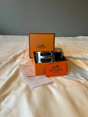 H@rmes bälte - Säljer ett svart läderbälte med det ikoniska H-spännet i silverfärg. Bältet har vita kontrastsömmar och kommer i original orange H-box med tillhörande certifikat och kvitto. Perfekt accessoar för att lyfta vilken outfit som helst.