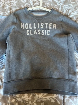 Grå sweatshirt från Hollister - Mysig grå sweatshirt från Hollister med texten 'HOLLISTER CLASSIC' i vitt framtill. Tröjan har rund halsringning, ribbade muddar och är tillverkad i mjukt sweatshirtmaterial. Herr modell men fin på tjejer också. XLmen är mer som en M/L