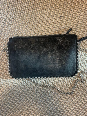 Söt handväska med löstagbar kedja - Cool svart handväska i mjukt skinn med silverfärgad kedja och metallkedjedetaljer runt kanterna. Väskan har en rektangulär form och passar perfekt för att bära det viktigaste. Snygg och enkel design som ger en edgy look.