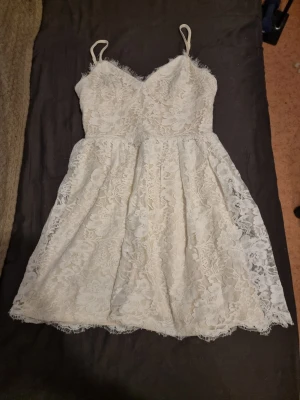Vit spetsklänning med smala axelband - Säljer en söt vit kortklänning med spetsdetaljer och smala axelband. Klänningen är kort, har en v-ringad överdel och är ärmlös, perfekt för sommarens alla tillfällen. Den har en lätt utsvängd kjol och romantisk känsla.