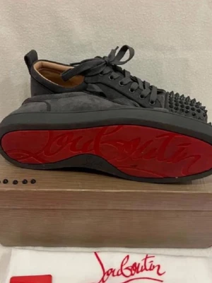 Sneakers från Louboutin med nitar - Säljer ett par sneakers från Christian Louboutin med klassisk röd sula och nitar på tån. Skorna har snörning och är tillverkade i mocka för en lyxig känsla. Perfekta för dig som vill sticka ut med en edgy detalj.