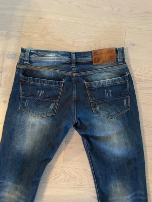 Niche Diesel jeans - Säljer ett par blå jeans från Diesel med snyggt sliten tvätt och detaljerade bakfickor. Jeansen har klassisk femficksmodell, raka ben och en skinnpatch med Diesel-logga bak i midjan. Materialet är denim i bomull med lite stretch för extra komfort. Modellen är 188cm