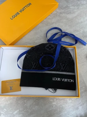 L,V mössa - Snygg svart mössa från HM med ikoniskt monogrammönster och grå rand. Märkets logga broderad framtill. Kommer i originalförpackning med tillhörande band och kort. Perfekt accessoar för att lyfta din stil. Är knappt använd och måsarna i perfekt kondition.