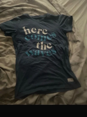 Blå t-shirt med tryck 'here comes the waves' - Mörkblå t-shirt med vit och ljusblå text på framsidan där det står 'here comes the waves'. Klassisk passform med rund hals och korta ärmar. Perfekt för en avslappnad stil.