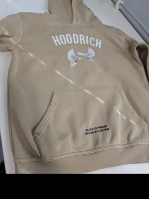 Beige hoodie från Hoodrich XL - Snygg beige hoodie från Hoodrich med vit text och en grafisk duva på bröstet. Stor känguruficka framtill och trycket 'ALWAYS PROUD NEVER SATISFIED' på fickan. Mjuk insida och klassisk huva, perfekt för en chill streetwear-look.
