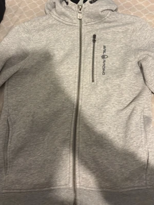 Ljusgrå hoodie från Sail Racing - Ljusgrå hoodie från Sail Racing med dragkedja framtill, huva och en bröstficka med dragkedja. Tillverkad i mjukt material och har logga tryckt på bröstet. Perfekt för en avslappnad och sportig stil.