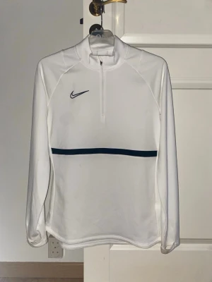 Vit träningströja Nike  - Vit långärmad träningströja från Nike med halv dragkedja framtill och svart logga på bröstet. Tröjan har tumhål och är tillverkad i ett mjukt, stretchigt funktionsmaterial. Perfekt för träning eller sportaktiviteter. Storlek S fint skick använd ett par gånger bara