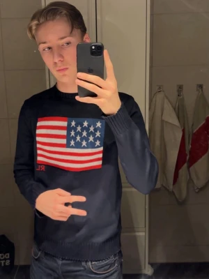 Mörkblå stickad Ralph lauren tröja med USA-flagga - Snygg mörkblå stickad tröja från Ralph Lauren med ett stort USA-flagga motiv på bröstet och RL-broderi. Tröjan har rund hals och ribbade muddar vid ärmslut och nederkant. Perfekt för dig som gillar klassisk stil.