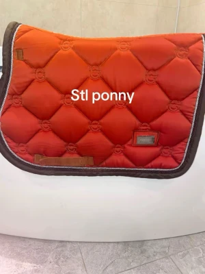 Orange schabrak ponny Equestrian Stockholm - Säljer ett snyggt orange schabrak från Equestrian Stockholm i ponny-storlek. Schabraket har bruna kanter, quiltat mönster och en metallbricka med logga. Perfekt för dig som vill sticka ut i stallet!