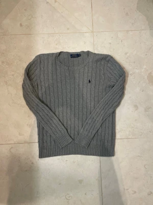 Grå kabelstickad tröja från Polo Ralph Lauren - Snygg grå kabelstickad tröja från Polo Ralph Lauren med rund hals och diskret broderad logotyp på bröstet. Tröjan har långa ärmar och ribbade muddar vid ärmslut och nederkant. Perfekt för lager-på-lager och en klassisk look.står M men passar mer en S 