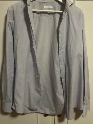 Ljusblå skjorta från J.Lindeberg - Klassisk ljusblå skjorta från J.Lindeberg med lång ärm och knappar framtill. Skjortan har en enkel, stilren design och är tillverkad i mjuk bomull som känns skön mot huden. Perfekt för dig som gillar en clean och tidlös look.