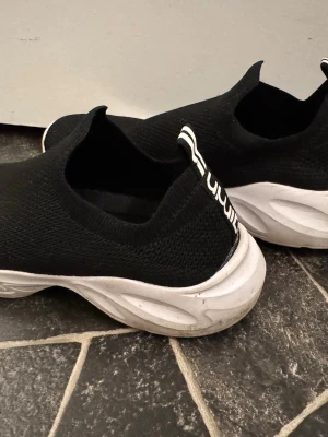 Svarta slip-on sneakers med vit sula - Säljer ett par svarta slip-on sneakers med stickad ovandel och chunky vit sula. Skorna har en elastisk passform och en svart/vit flärp bak för enkel påtagning. Perfekta för dig som gillar en clean och sportig look.