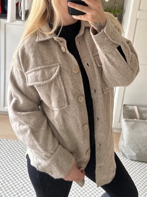 Beige overshirt med fickor - Stilren beige jacka i mjukt, strukturerat tyg med stora knappar framtill och två bröstfickor med lock. Rak passform och klassisk krage, perfekt att slänga över en hoodie eller t-shirt för en avslappnad look.