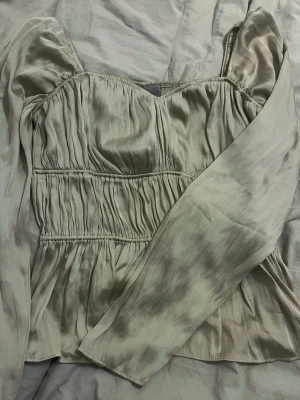 Beige satinblus med puffärm - Trendig beige blus i glansigt satinmaterial med smockdetaljer och långa puffärmar. Snyggt rynkad över bysten och midjan för en feminin siluett. Perfekt att styla med jeans eller kjol för en chic look.