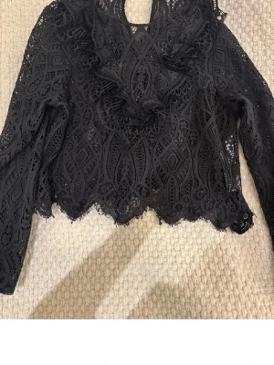 Svart spetsblus med volanger - Säljer en svart spetsblus med långa ärmar och snygga volangdetaljer framtill. Blusen har ett genombrutet mönster och vågig kant nertill, vilket ger en trendig och elegant look. Perfekt att styla med jeans eller kjol för en cool outfit.