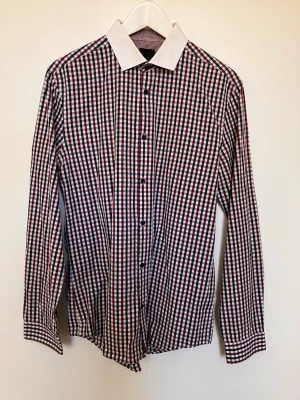 Rutig skjorta med vit krage  - Snygg långärmad H&M skjorta med vinrött, blått och vitt rutmönster samt kontrasterande vit krage. Klassisk passform och knäppning framtill. Perfekt för dig som vill ha en stilren men ändå unik look. Säljes i befintligt skick enligt bilderna!