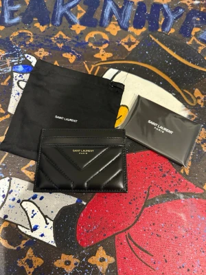 Svart kortfodral från Saint Laurent - Snyggt svart kortfodral från Saint Laurent i glansigt läder med chevronmönster och guldfärgad logga framtill. Perfekt för att hålla koll på dina kort med stil. Kommer med originalpåse och kort.