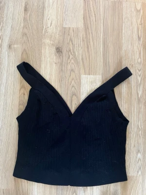 Svart croppad linnetopp - Trendig svart croppad linnetopp med breda axelband och djup v-ringning. Toppen har en ribbad struktur som ger en snygg detalj och passar perfekt till höga jeans eller kjol. Enkel och stilren design som är lätt att matcha.