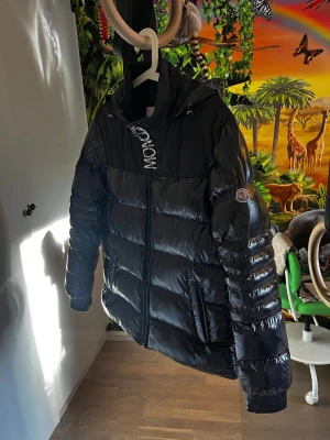 Svart dunjacka  - Snygg svart dunjacka  med glansig finish och klassisk logga på ärmen. Jackan har huva, dragkedja framtill och breda kanaler fyllda med dun. Perfekt för kalla vinterdagar och riktigt trendig stil.