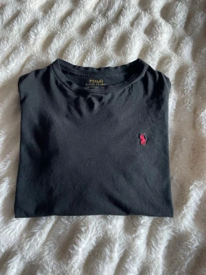 Svart t-shirt från Polo Ralph Lauren - Svart t-shirt från Polo Ralph Lauren med klassisk passform och korta ärmar. Ikoniska röda Polo-loggan broderad på bröstet. Tillverkad i 100% bomull för en skön och mjuk känsla. Perfekt basic-plagg med stilren look.