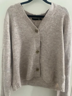 Beige kofta från 157 med knappar - Mysig beige kofta från 157 med v-ringning och tre stora knappar framtill. Koftan har lång ärm och är stickad i ett mjukt material som känns skönt mot huden. Perfekt för lager på lager och enkel att matcha med olika outfits.