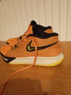 Nike Kyrie orange basketskor - Säljer ett par Nike Kyrie basketskor i orange med svarta detaljer och gula inslag. Skorna har en låg profil, svart swoosh på sidan och meshmaterial för bra ventilation. Sulan är vit med gul undersida och skorna har snörning framtill. Perfekta för basket eller streetwear.