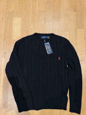 Svart stickad tröja Polo Ralph Lauren - Svart stickad tröja från Polo Ralph Lauren med rund halsringning och röd broderad logga på bröstet. Tröjan har ribbade muddar vid ärmslut och nederkant. Klassisk och stilren design som passar till mycket.
