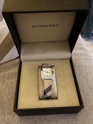 Burberry rutmönstrad klocka beige - Säljer en stilren damklocka från Burberry, modell BU1577. Klassisk rektangulär urtavla och det ikoniska rutiga Burberry-bandet.  Mycket fint skick och knappt använd, Swiss Made, har datumvisning och är vattentålig upp till 50m.   Originalbox medföljer. Kvittot finns tyvärr inte kvar, men alla originalnummer finns graverade på baksidan av klockan. 