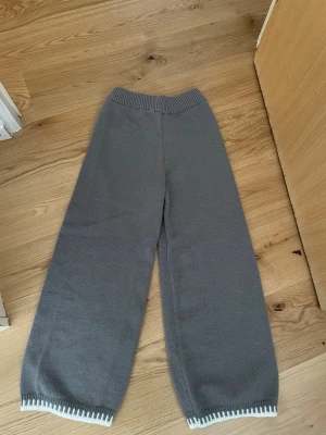 Grå stickade mjukisbyxor - Mysiga grå stickade mjukisbyxor med bred resår i midjan och raka, vida ben. Byxorna har vita detaljer längst ner vid bensluten som ger en snygg kontrast. Perfekta för chill dagar hemma eller när du vill vara extra bekväm.