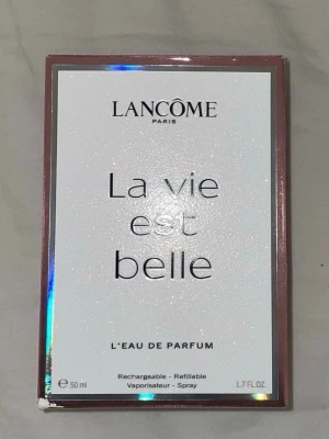 Lancôme La Vie Est Belle EdP 50ml - Lancôme La Vie Est Belle Eau de Parfum i 50 ml flaska. Parfymen kommer i en elegant, rektangulär glasflaska med persikofärgad vätska och en dekorativ rosett vid halsen. Förpackningen är vit med skimrande detaljer och roséguldram.
