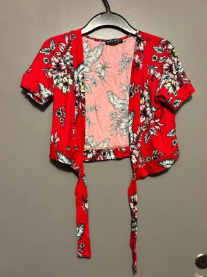 Röd blommig omlottopp från Select - Snygg röd omlottopp från Select med blommigt mönster i vitt, grönt och svart. Toppen har korta ärmar och knyt framtill för en trendig look. Perfekt för dig som vill sticka ut med färg och mönster.
