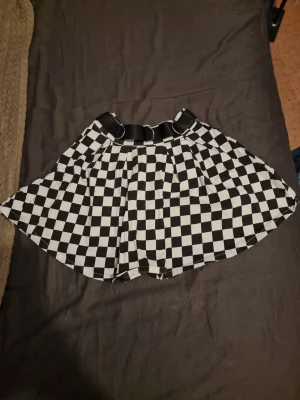 Rutig svartvit kortkjol med bälte - Säljer en svartvit kortkjol med schack rutigt mönster och ett brett bälte med metallringar i midjan. Kjolen har en cool streetstyle-vibe och passar perfekt till sneakers eller boots. Materialet känns lätt och bekvämt, perfekt för dig som vill sticka ut.