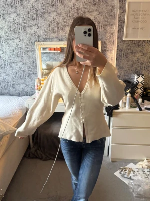 Krämvit kofta med v-ringning - Snygg krämvit kofta med v-ringning och knappar framtill. Koftan har långa, lite puffiga ärmar och en lätt figursydd passform. Perfekt att slänga över en topp för en stilren look.