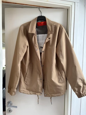 Beige vindjacka från Selected Homme (S/M) - Stilren beige vindjacka från Selected Homme, modell The Coach. Jackan har tryckknappar, två snedställda fickor, dragsko i nederkant och elastiska muddar vid ärmsluten. Insidan har en innerficka och detaljer i grått. Perfekt för blåsiga dagar.