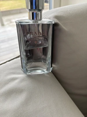 Prada L'Homme Eau de Toilette - Säljer en lyxig parfym från Prada, L'Homme. Flaskan är stilren i genomskinligt glas med silverfärgad metallkork och svart detalj på sidan. Innehållet är lila. Perfekt för dig som gillar exklusiva dofter och snygg design. Ca 65% kvar.