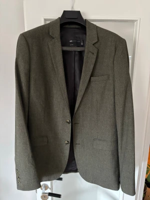 Olivgrön kavaj från ASOS design - Stilren olivgrön kavaj från ASOS design med två knappar framtill, klassisk krage och tre fickor. Kavajen har mönstrad yta och är helfodrad med svart innerfoder. Passar perfekt till en dressad look och har en tidlös design.