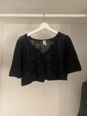 Svart kortärmad stickad kofta - Snygg svart stickad kofta med korta ärmar och croppad passform. Koftan har ett hålmönster och knäppning framtill. Perfekt att slänga över ett linne eller klänning. Det står storlek M men passar även min som brukar ha S. Köpt för 350kr men säljs för 70kr.