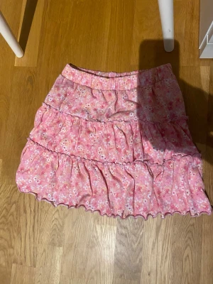 Rosa blommig volangkjol med shorts inuti  - Supersöt kort kjol i rosa med små blommor och volangdetaljer. Kjolen har resår i midjan och är gjord i ett lätt, mjukt tyg som passar perfekt till varma dagar. Perfekt för dig som gillar en romantisk och lekfull stil.