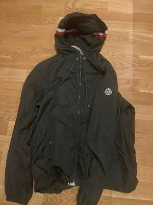 Mörkgrön Moncler vindjacka med huva - Svart vindjacka från Moncler med huva och dragkedja framtill. Jackan har Moncler-logga på bröstet och röda/vita detaljer på huvan. Materialet är lätt och vattenavvisande, perfekt för blåsiga dagar. Två fickor med tryckknappar och justerbar dragsko i huvan.