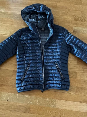 Colmar Mörkgrå pufferjacka med huva - Snygg mörkgrå pufferjacka från Colmar med quiltad design och fast huva. Jackan har två dragkedjeförsedda fickor framtill och en logotyp på ärmen. Storlek 48 Italien storlek motsvarar 40/42 