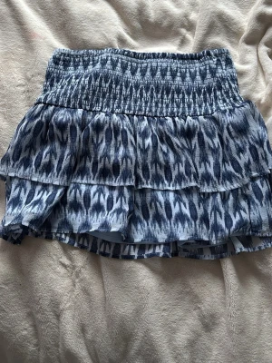 Blå mönstrad volangkjol Gina Tricot - Kort volangkjol från Gina Tricot i blått och vitt med elastisk smock i midjan. Kjolen har ett snyggt abstrakt mönster och dubbla volanger som ger en luftig och trendig look. Perfekt för dig som gillar en avslappnad men ändå stilsäker outfit.