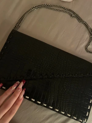 Svart axelväska med kedjerem - Snygg svart axelväska i fejkläder med croco-mönster och silverfärgad kedjerem. Väskan har en kuvertformad design och dekorativa sömmar längs kanterna. Perfekt för dig som gillar trendiga accessoarer med lite edge.
